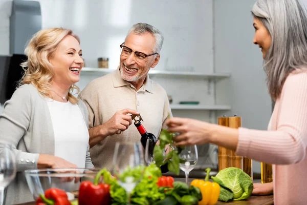 Donna sorridente che cucina e parla con un amico, uomo che apre bottiglia con vino — Foto stock