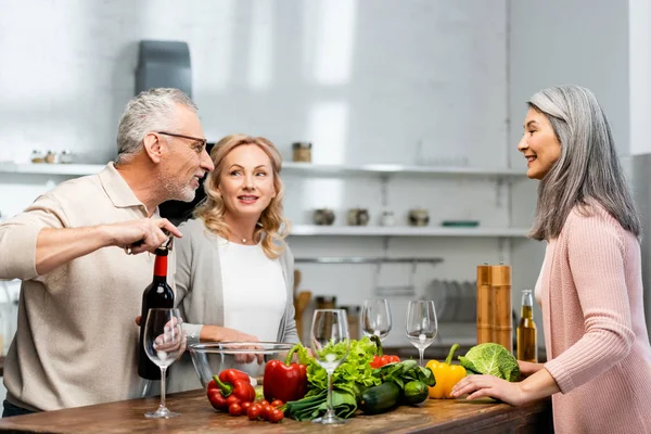 Mulher sorrindo cozinhar, homem abrindo garrafa com vinho e falando com mulher asiática — Fotografia de Stock