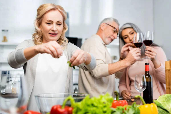 Enfoque selectivo de la mujer agregando lechuga al tazón y sus amigos multiculturales mirando copas de vino en el fondo - foto de stock