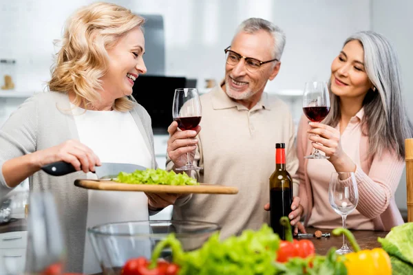 Sonriente mujer sosteniendo tabla de cortar con lechuga y sus amigos multiculturales sosteniendo copas de vino - foto de stock