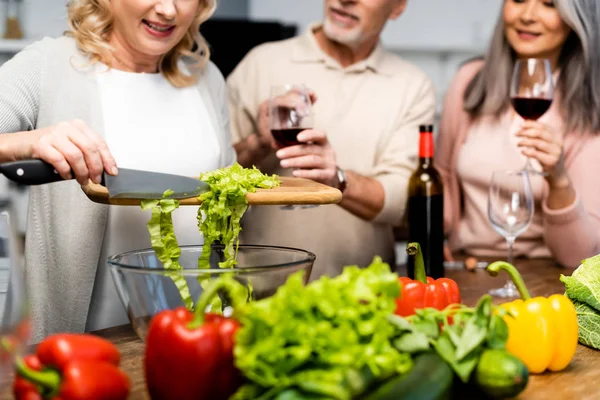 Обрезанный вид женщины, добавляющей салат в миску и ее друзей, держащих бокалы для вина — стоковое фото