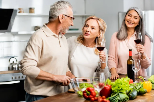 Smiling multicultural woman holding wine glasses and man cutting lettuce in kitchen — Stock Photo