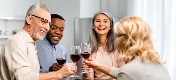 Panoramic shot of smiling multicultural friends clinking with wine glasses in kitchen — Stock Photo