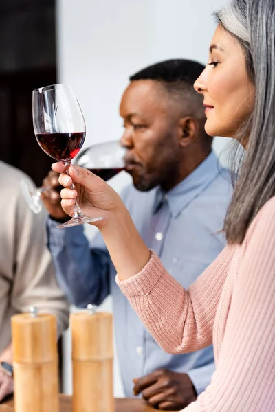 Foco seletivo de mulher asiática olhando para o copo de vinho e homem americano africano bebendo vinho — Fotografia de Stock
