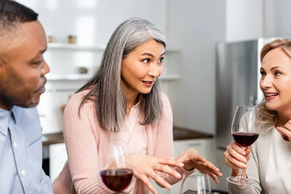 Enfoque selectivo de sonreír amigos multiculturales hablando y sosteniendo copas de vino en la cocina - foto de stock