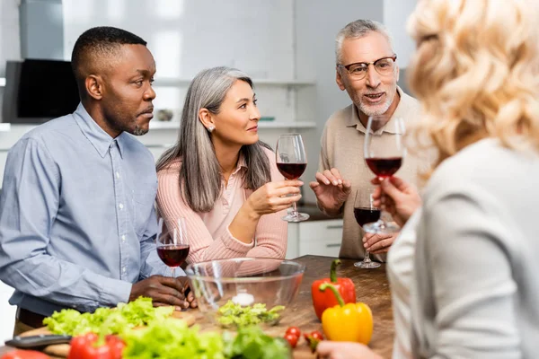 Foco seletivo de amigos multiculturais falando e segurando copos de vinho na cozinha — Fotografia de Stock
