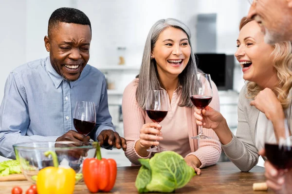 Enfoque selectivo de sonreír amigos multiculturales hablando y sosteniendo copas de vino en la cocina - foto de stock