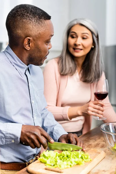 Africano americano hombre corte lechuga y hablando con asiático amigo - foto de stock