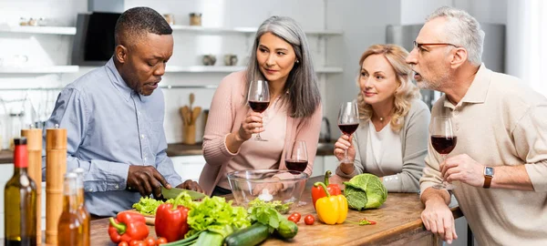 Scatto panoramico dell'uomo afroamericano che taglia lattuga e parla con amici multiculturali — Foto stock