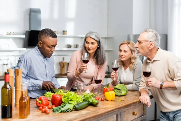 Uomo afroamericano taglio lattuga e parlare con amici multiculturali — Foto stock