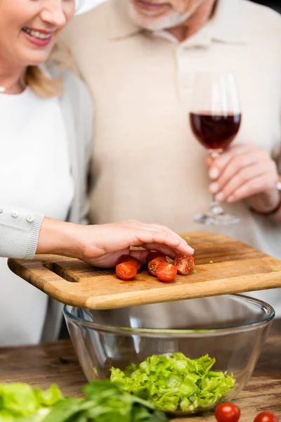 Обрезанный вид улыбающейся женщины, добавляющей вырезанные помидоры черри в миску, и мужчина, держащий бокал вина — стоковое фото