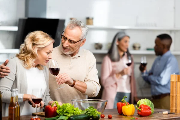 Foco seletivo de homem sorridente segurando copo de vinho e mulher cheirando, amigos multiculturais falando em segundo plano — Fotografia de Stock