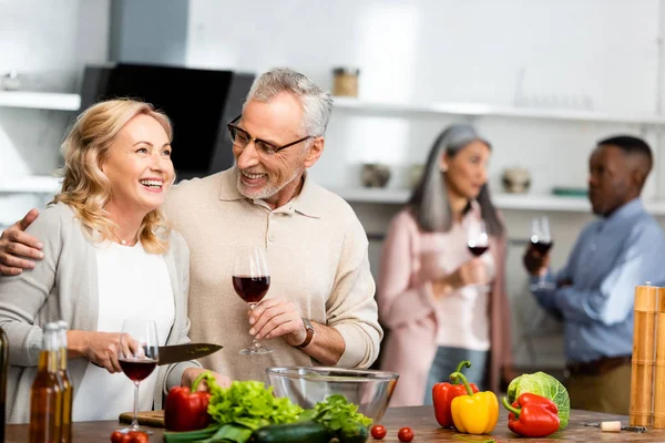 Enfoque selectivo de hombre sonriente sosteniendo copa de vino y mujer sosteniendo cuchillo, amigos multiculturales hablando sobre el fondo - foto de stock