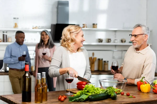 Selective focus of woman cutting bell pepper and talking with man, multicultural friends talking on background — Stock Photo