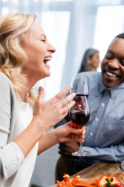 Избирательный фокус улыбающейся женщины и африканского американца, говорящего и держащего бокалы для вина — стоковое фото