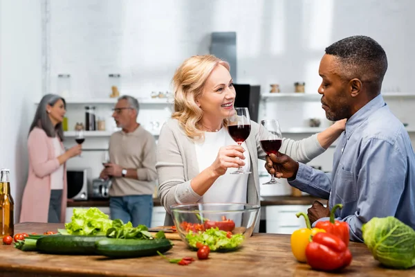 Selective focus of smiling woman and african american man talking and holding wine glasses, multicultural friends talking on background — Stock Photo