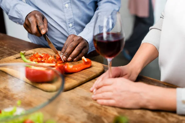 Vista cortada de homem afro-americano cortando pimentão e mulher segurando copo de vinho — Fotografia de Stock