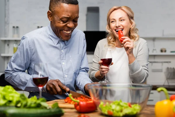 Souriant homme afro-américain coupe poivron et femme tenant verre de vin et manger poivron — Photo de stock