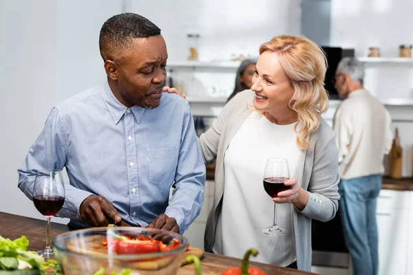 Афроамериканський чоловік ріже болгарський перець і жінка тримає келих для вина і розмовляє з ним — стокове фото