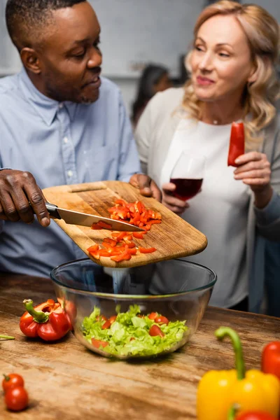 Messa a fuoco selettiva di uomo afro-americano sorridente aggiungendo taglio peperone a ciotola e donna in possesso di bicchiere di vino e peperone — Foto stock