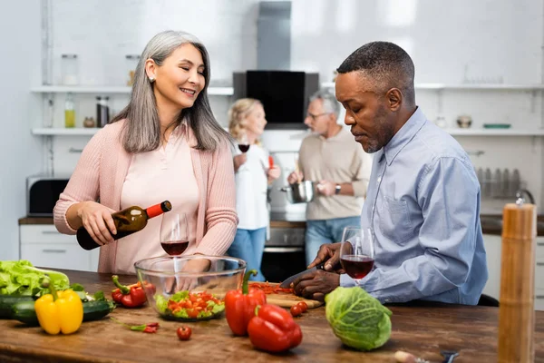 Foco seletivo de sorrir asiático mulher derramando vinho para vidro e africano americano homem corte sino pimenta — Fotografia de Stock