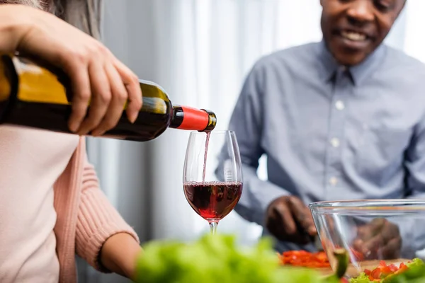 Vista cortada de mulher derramando vinho para vidro e homem americano africano olhando para ele — Fotografia de Stock