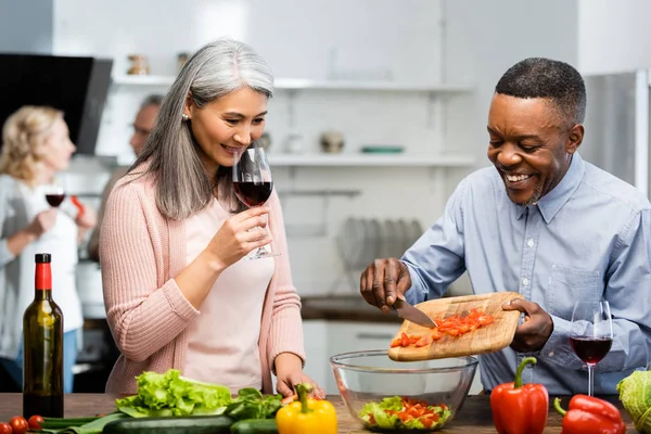 Smiling african american man adding cut bell pepper to bowl and asian woman smelling wine — Stock Photo