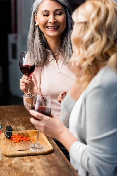 Избирательный фокус улыбающейся азиатской женщины, разговаривающей с другом и держащей бокал вина — стоковое фото