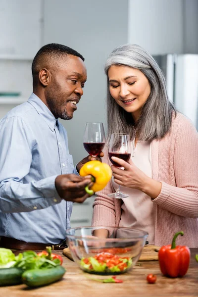 Africano americano hombre holding campana pimienta y hablando con sonriente asiático amigo - foto de stock