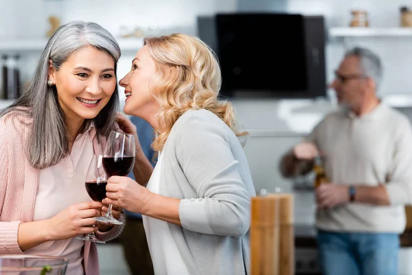 Selettivo fuoco di sorridente donna dicendo segreto per asiatico amico con vino bicchiere — Foto stock