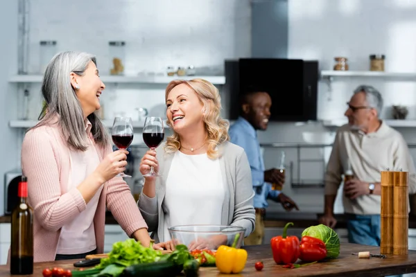 Selective focus of smiling multicultural friends holding wine glasses in kitchen — Stock Photo