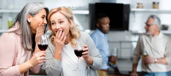 Panoramic shot of asian woman telling secret to shocked friend and multiethnic friends on background — Stock Photo