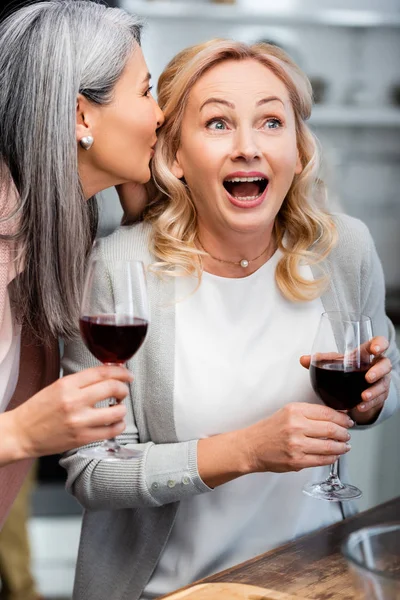 Asiático mujer diciendo secreto a impactado amigo con vino vidrio - foto de stock