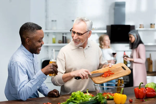 Afro-americano segurando cerveja e seu amigo adicionando tomate cereja à salada — Fotografia de Stock