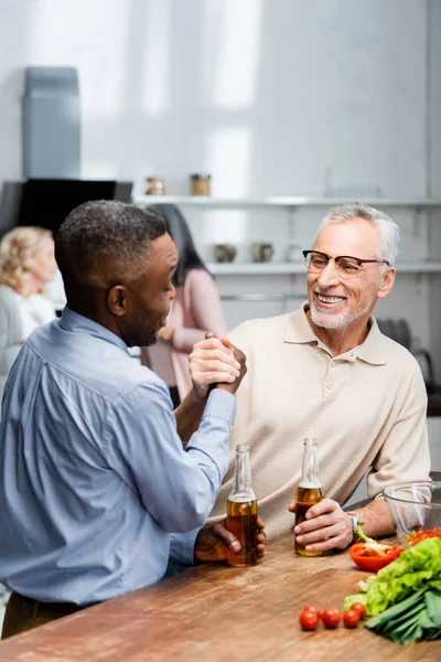 Africano americano hombre cogido de la mano y hablando con sonriente amigo en cocina - foto de stock