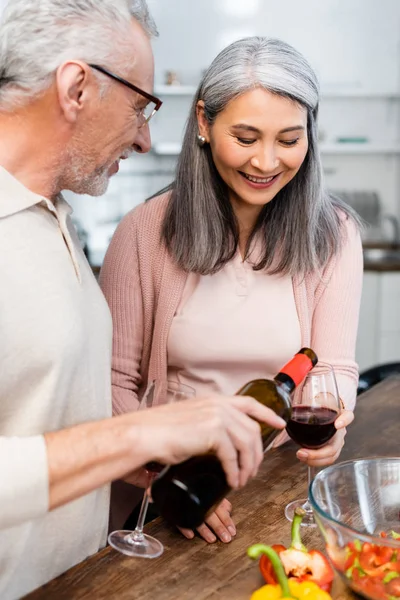 Smiling man pouring wine for his asian friend in kitchen — Stock Photo