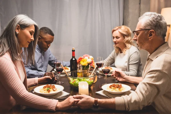 Amigos multiculturales tomados de la mano y rezando durante la cena - foto de stock