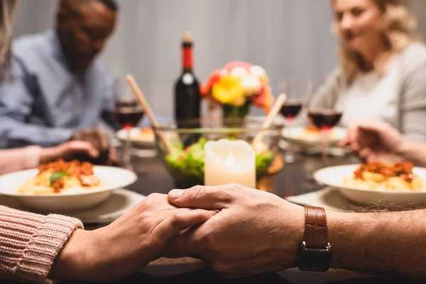 Vista cortada de amigos multiculturais de mãos dadas e rezando durante o jantar — Fotografia de Stock