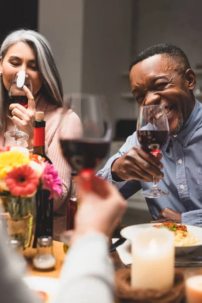 Selective focus of smiling asian woman and african american man holding wine glasses during dinner — Stock Photo