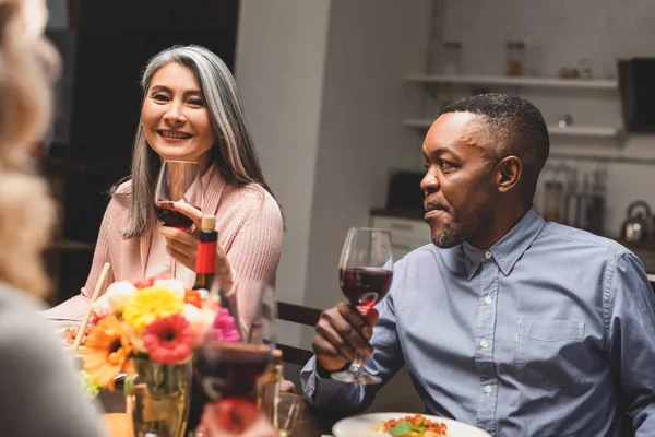 Foco seletivo de mulher asiática sorridente e homem americano africano segurando copos de vinho durante o jantar — Fotografia de Stock