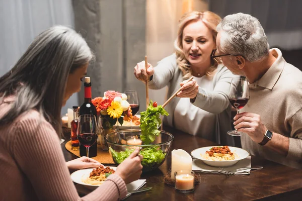 Lächelnde Frau nimmt Salat aus Schüssel und unterhält sich beim Abendessen mit Freundin — Stockfoto