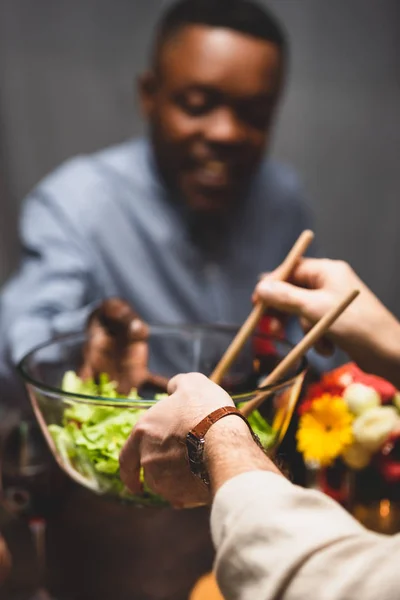 Vista cortada de mulher dando tigela com salada para amigo afro-americano durante o jantar — Fotografia de Stock