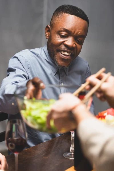 Cropped view of woman giving owl with salad to african american friend during dinner — Stock Photo