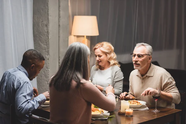 Selective focus of smiling man and woman talking with multicultural friends and eating during dinner — Stock Photo