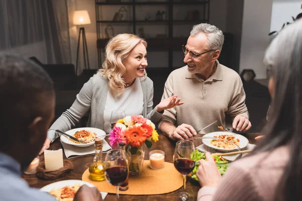 Selective focus of smiling man and woman talking with multicultural friends during dinner — Stock Photo