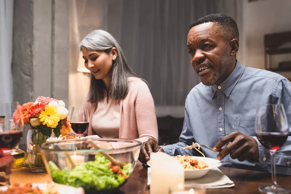 Amigos multiculturais sorridentes conversando e sentados à mesa durante o jantar — Fotografia de Stock