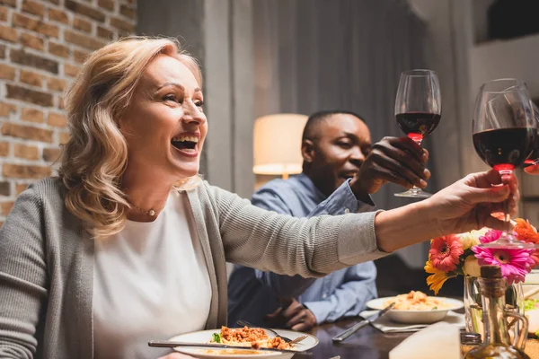 Selective focus of smiling multicultural friends clinking during dinner — Stock Photo