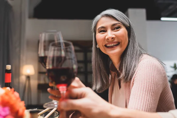 Sonriente y atractiva mujer asiática tintineo con amigo durante la cena - foto de stock