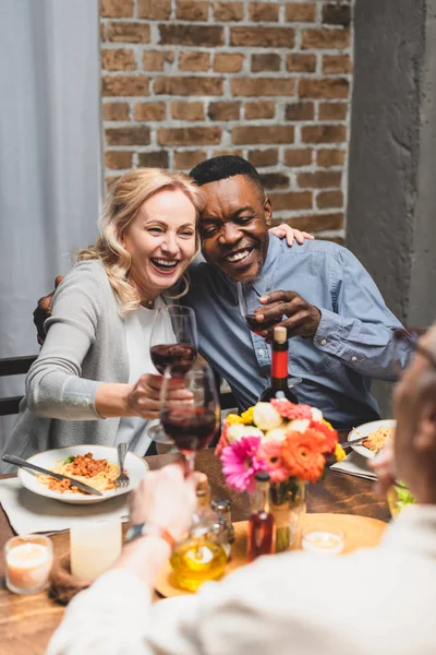 Enfoque selectivo de amigos multiculturales sonrientes abrazos y tintineo durante la cena - foto de stock