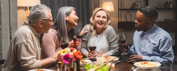 Tiro panorâmico de amigos multiculturais sorridentes falando e segurando copos de vinho durante o jantar — Fotografia de Stock